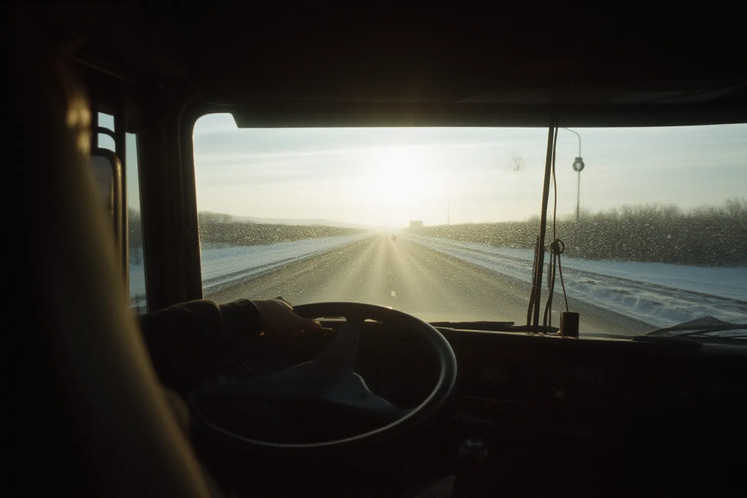 A hand on a large steering wheel inside a truck cab, low sun on a winter highway ahead